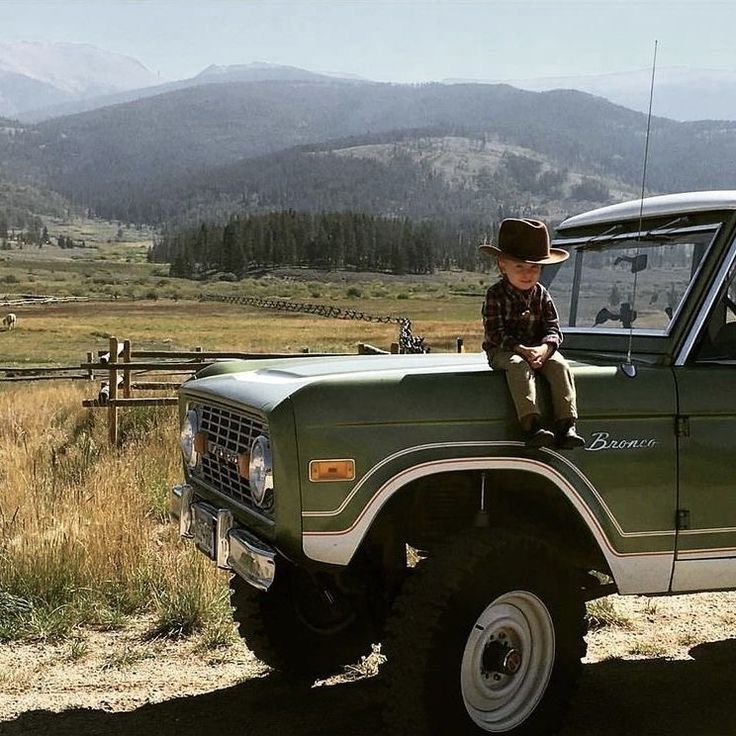 a young boy sitting on the hood of a green pick up truck with mountains in the background