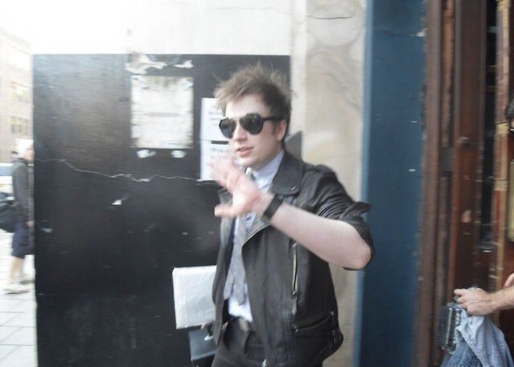 a man in sunglasses and a leather jacket standing on the sidewalk with his hand out