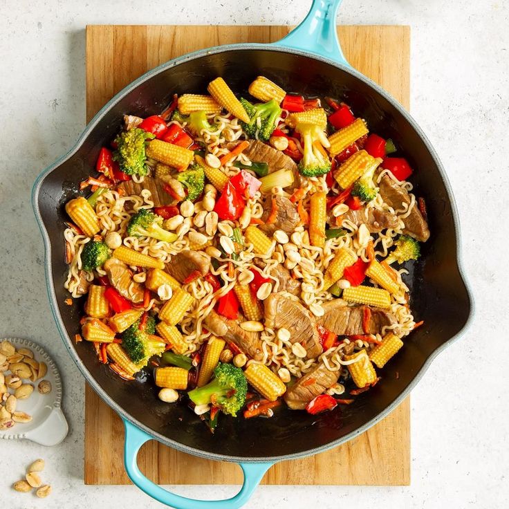 a skillet filled with pasta and vegetables on top of a wooden cutting board next to nuts