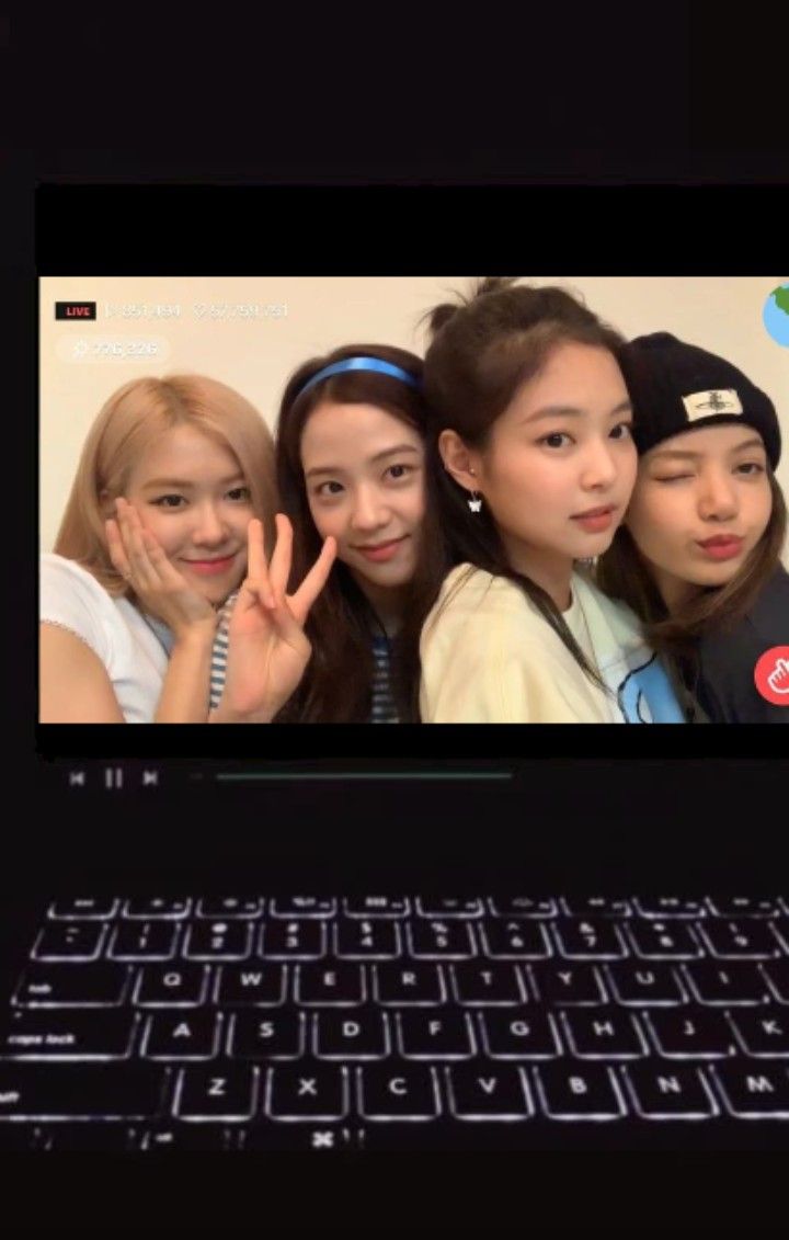 four girls are smiling and making the peace sign with their hands while standing in front of a laptop