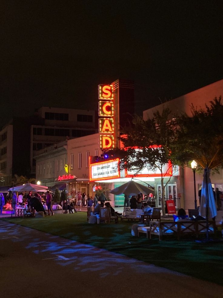 people are sitting at tables in front of a building with a neon sign on it