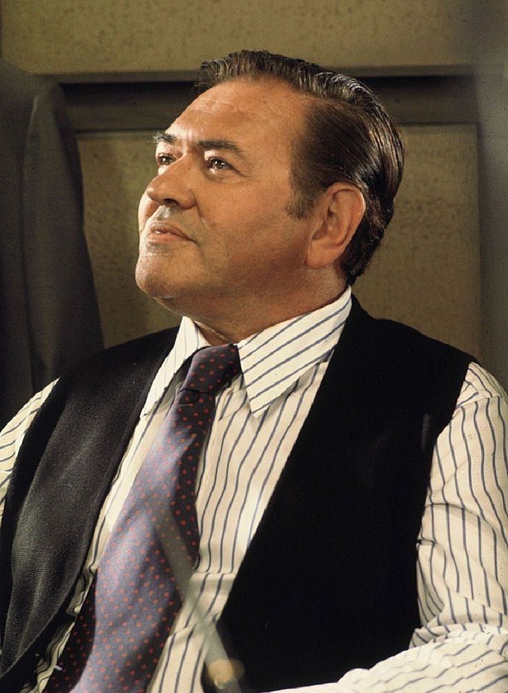 a man wearing a vest and tie sitting in a chair with his hand on his hip