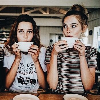 two young women sitting at a table drinking from coffee cups and looking up into the camera