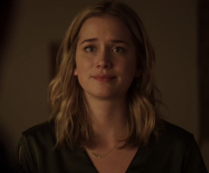 a woman with blonde hair looking at the camera in a dimly lit room, wearing a black shirt and gold necklace