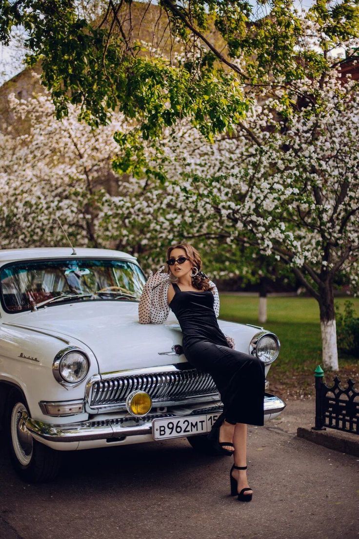 a woman sitting on the hood of an old fashioned car in front of some blossoming trees