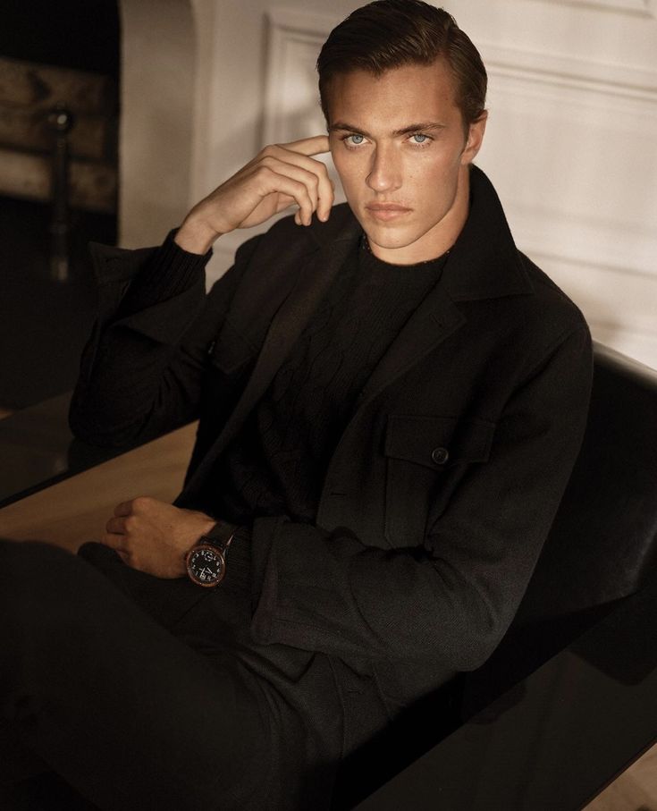 a man sitting on top of a wooden table wearing a black suit and watch in his hand