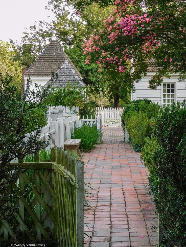 a brick path leading to a white house with pink flowers on the trees and bushes