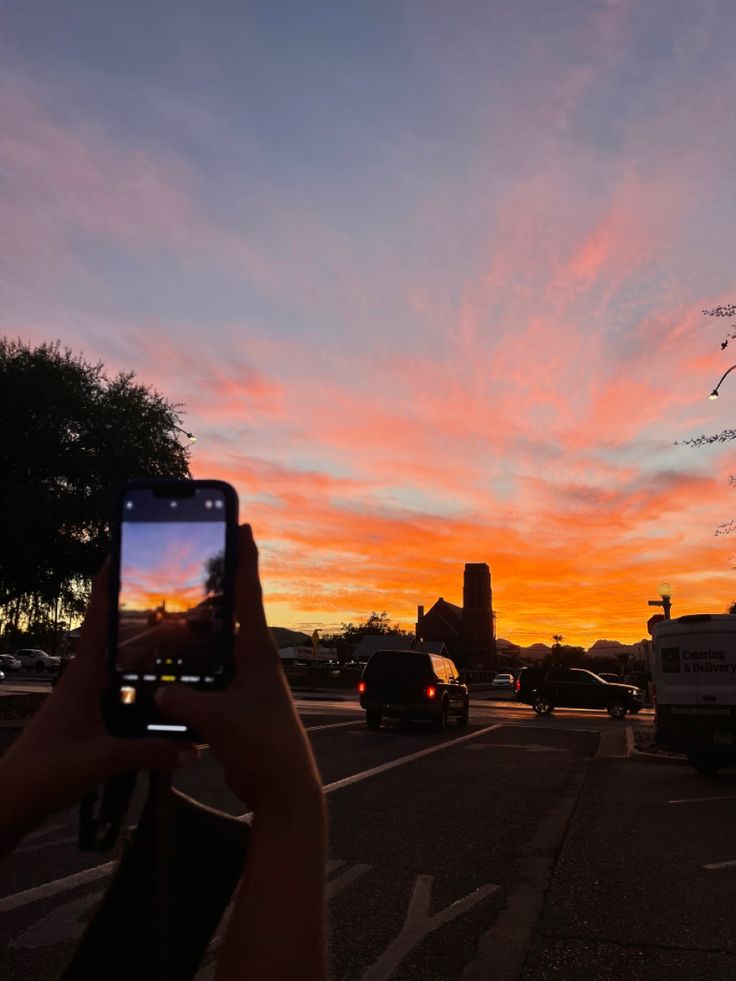 a person holding up a cell phone to take a photo of the sun setting over a parking lot