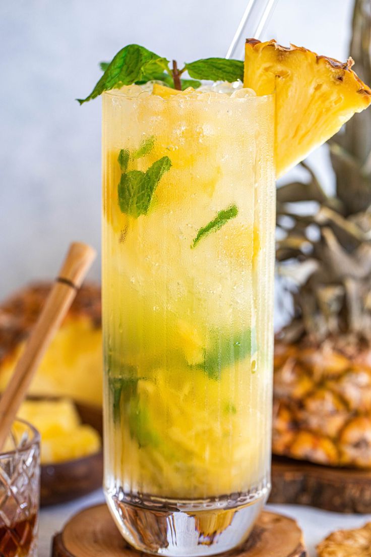 a pineapple drink with mint garnish in a tall glass next to sliced pineapples