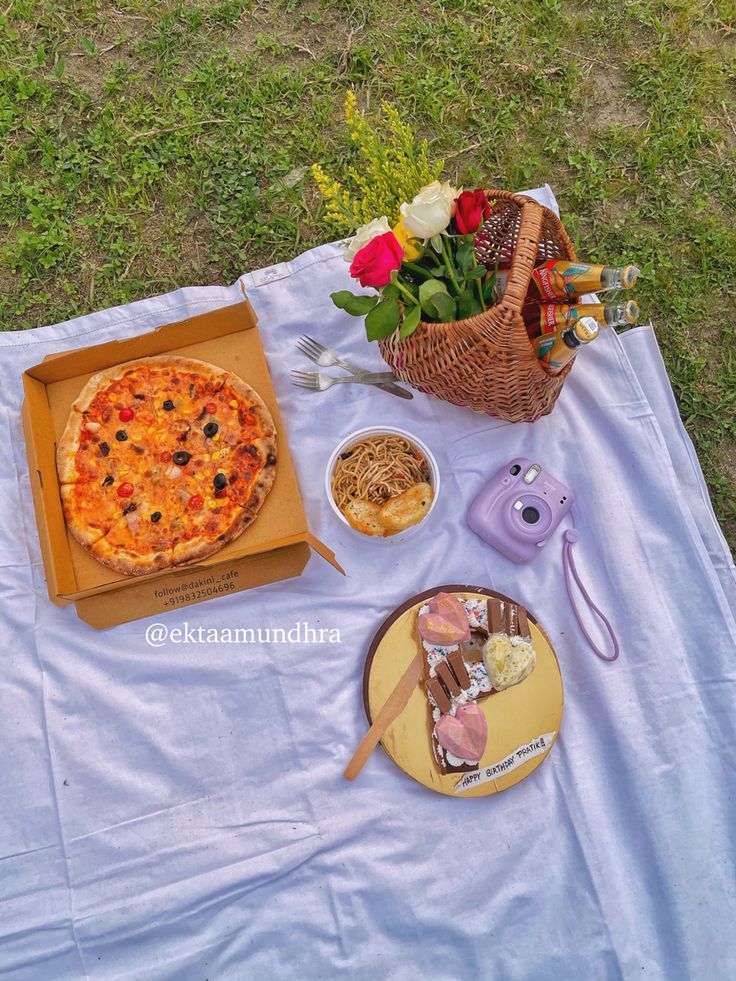 a pizza and other food items on a picnic blanket in the grass near some flowers