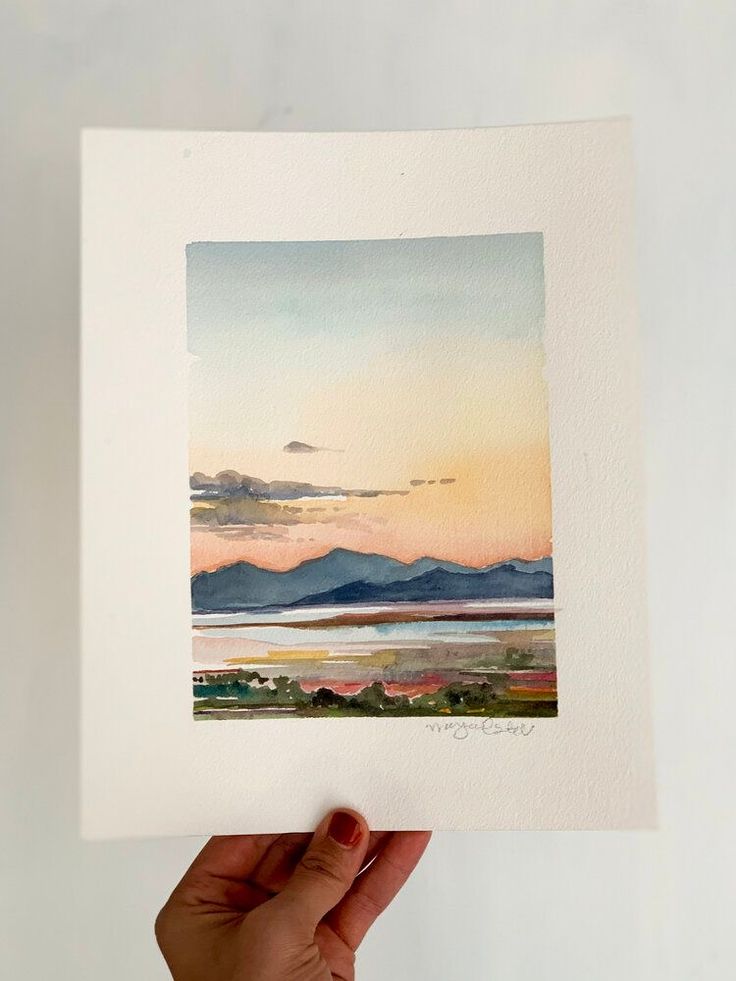 a hand holding up a watercolor painting with mountains in the background