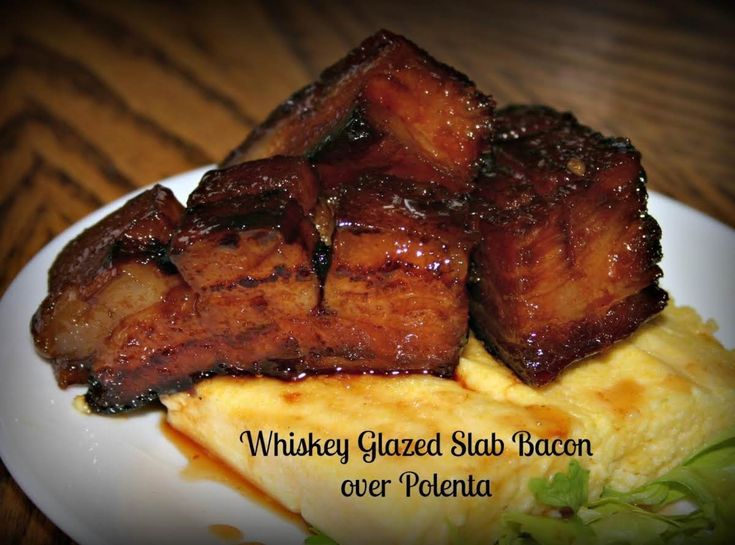 some food is on a white plate and has words that read whiskey glazed slab bacon over polenta