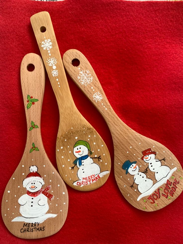 three wooden spoons decorated with snowmen on red fabric, one is for christmas and the other is for new year's eve