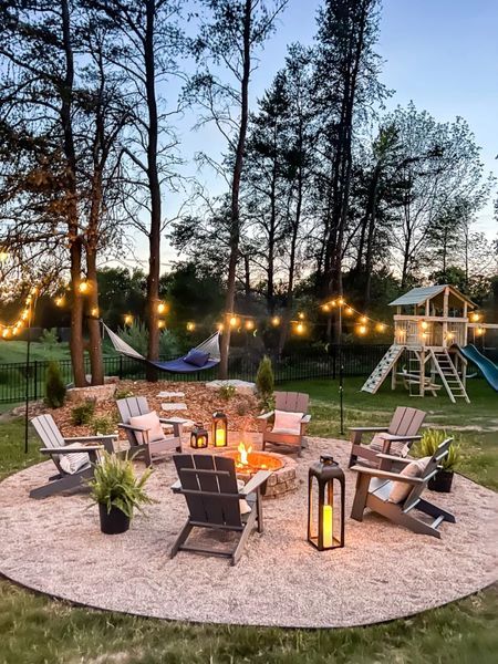 an outdoor fire pit surrounded by lawn chairs and lights in the evening with kids's play area behind it