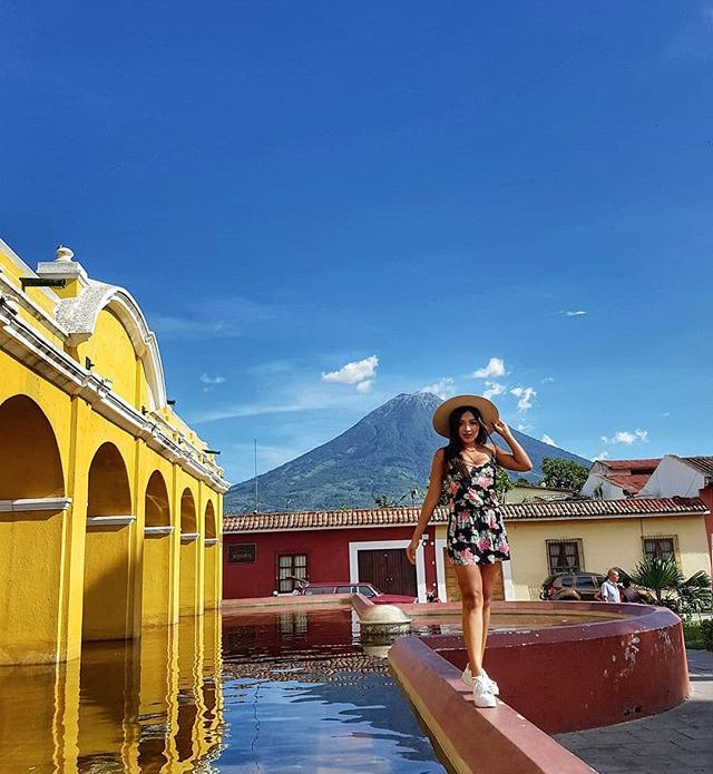 a woman wearing a hat standing in front of a fountain with mountains in the background