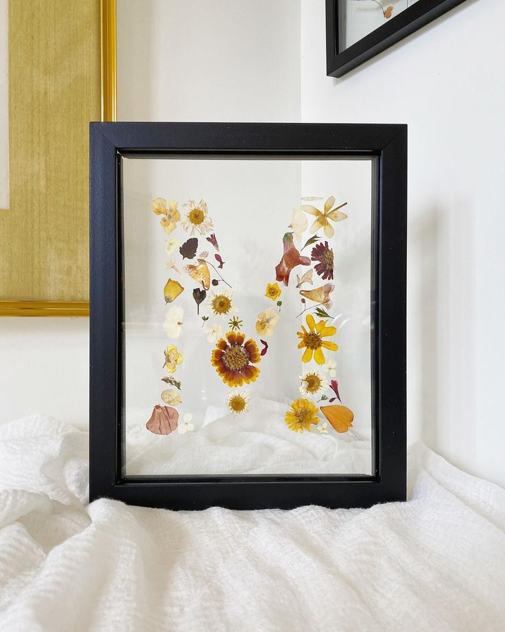 a framed letter made out of pressed flowers on a white bed with a gold frame