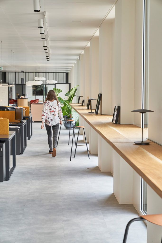 a woman walking down a long hallway between two desks with computers on them and plants in the background