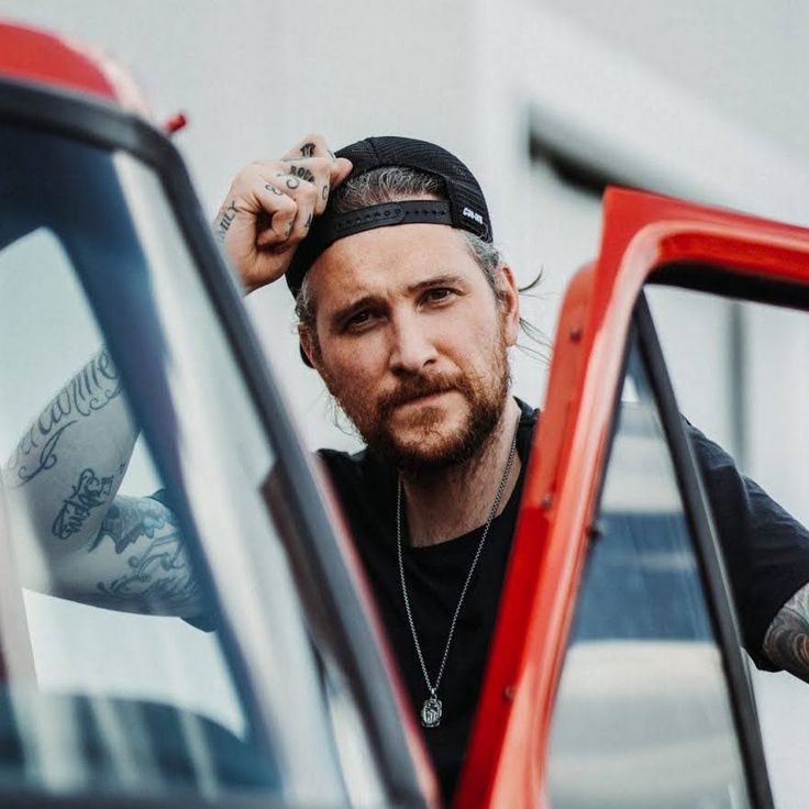 a man with tattoos leaning out the window of a red truck looking at the camera