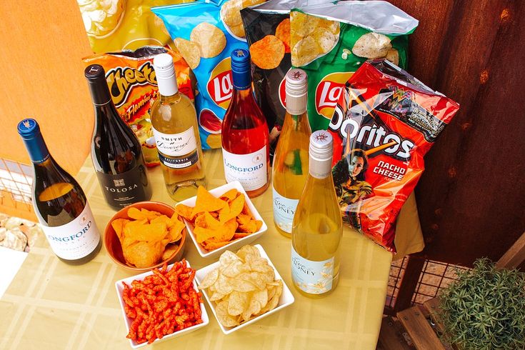 various snacks and drinks on a table with bottles of wine, crackers, chips, and popcorn