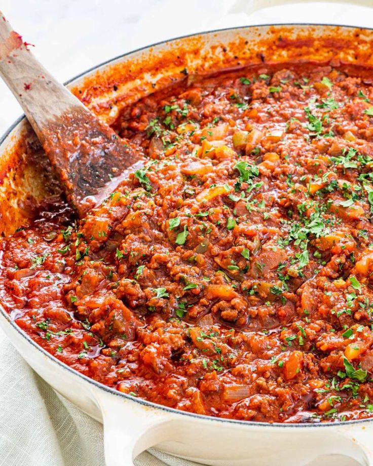 a large pot filled with meat and sauce on top of a white table cloth next to a wooden spoon