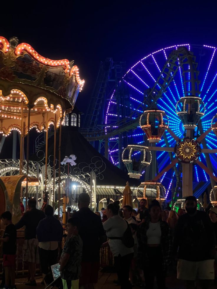 people are standing around in front of a ferris wheel at the fairground with lights on it