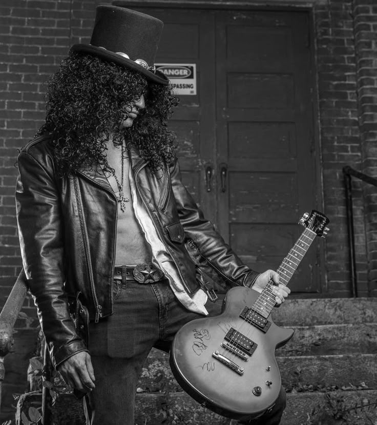 a man with long hair and a top hat holding a guitar in front of a brick building