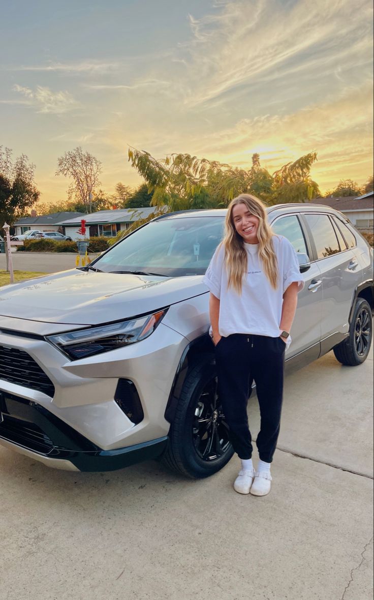 a woman standing in front of a silver car with the sun setting behind her and smiling