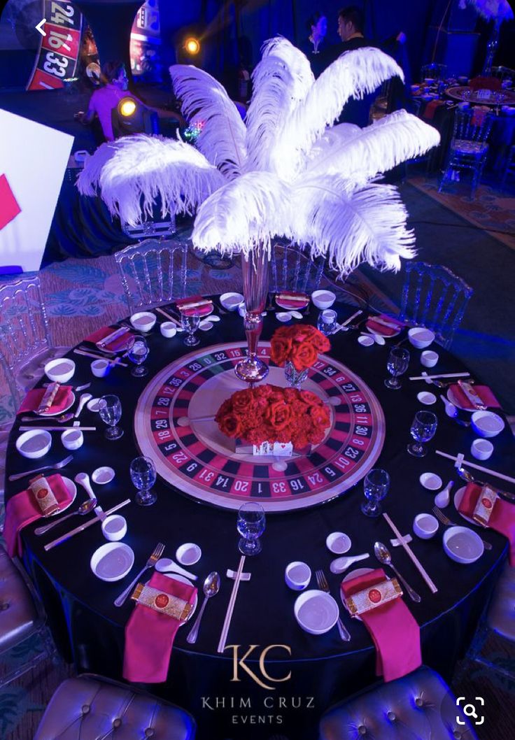 a casino roule with white feathers and red roses on the center table for an event