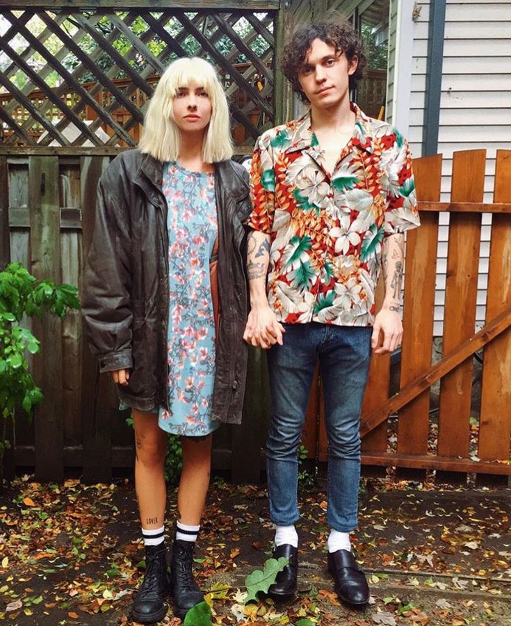 two people standing next to each other in front of a wooden fence with leaves on the ground