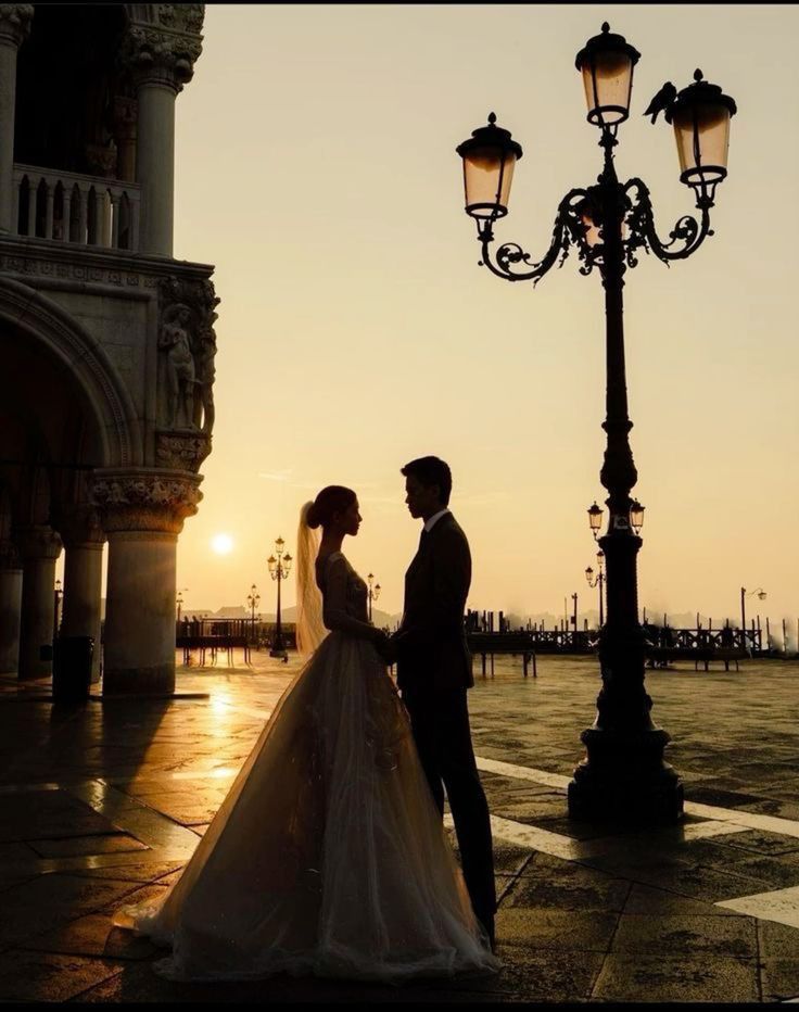 a bride and groom standing next to a street light in front of a building at sunset