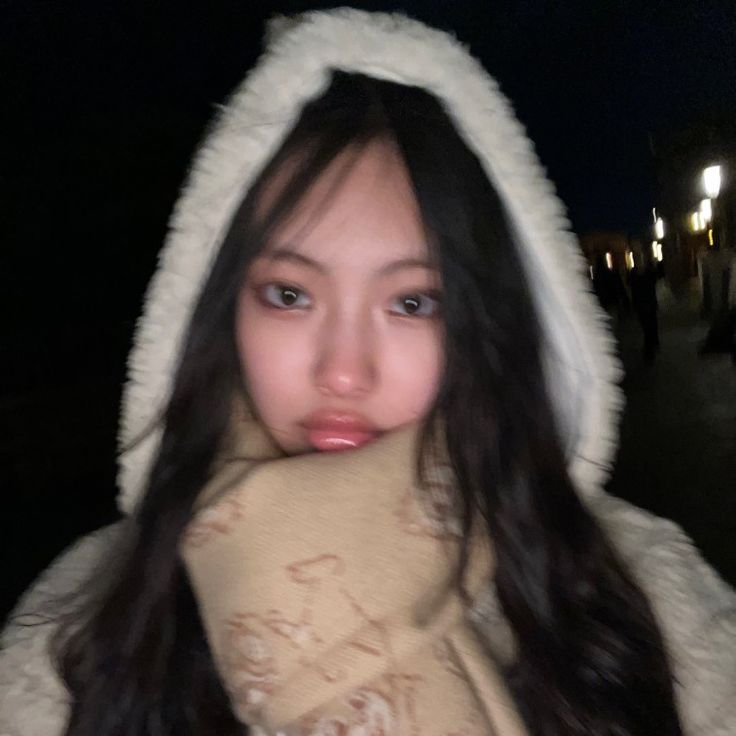 a woman with long black hair wearing a white coat and scarf on the street at night