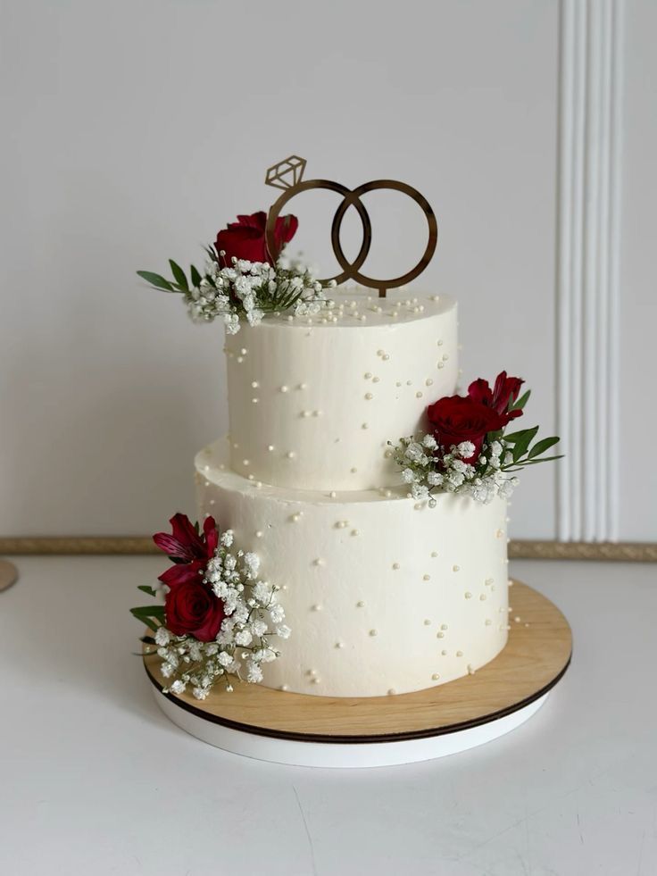 a three tiered wedding cake with red flowers and two rings on top, sitting on a wooden platter