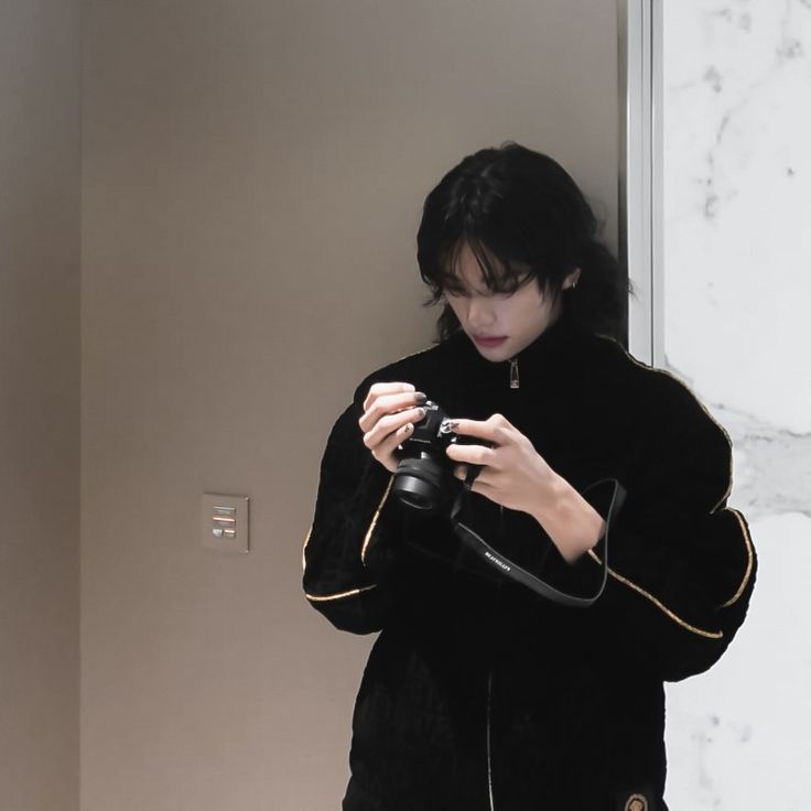 a woman is holding a camera and looking down at her phone while standing in front of a wall