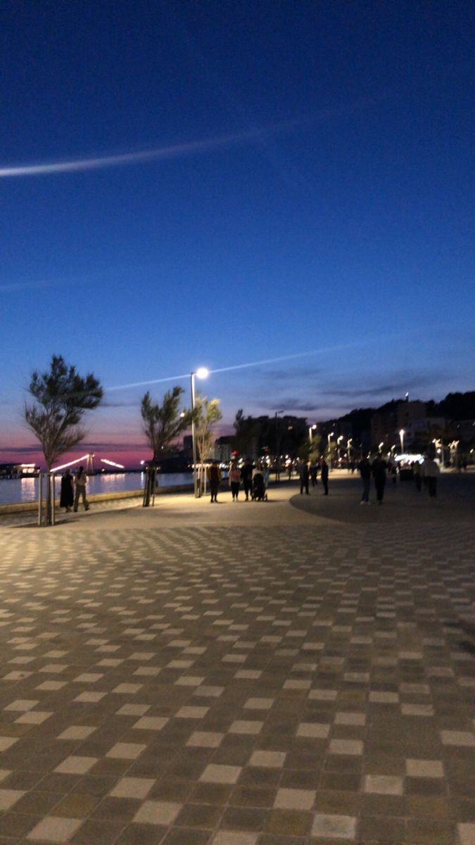 people are walking on the sidewalk at night near some water and buildings with lights in the background