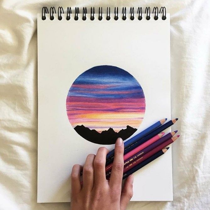 a person is drawing with colored pencils on top of a spiral notebook and there are mountains in the background