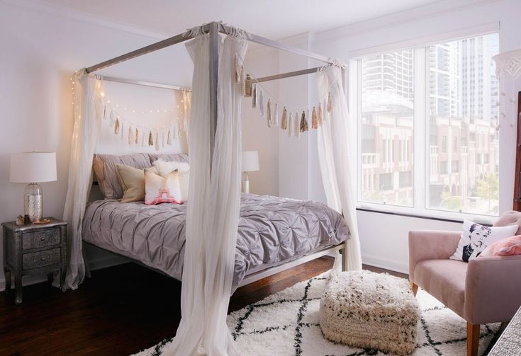 a white canopy bed sitting in a bedroom next to a pink chair and ottomans