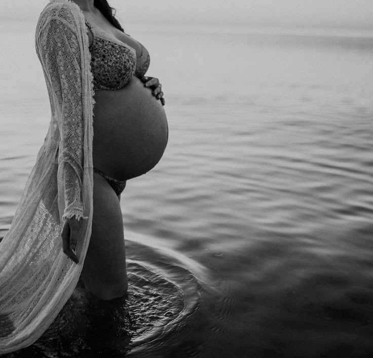 a pregnant woman standing in the water with her arms around her waist and wearing a sheer shawl