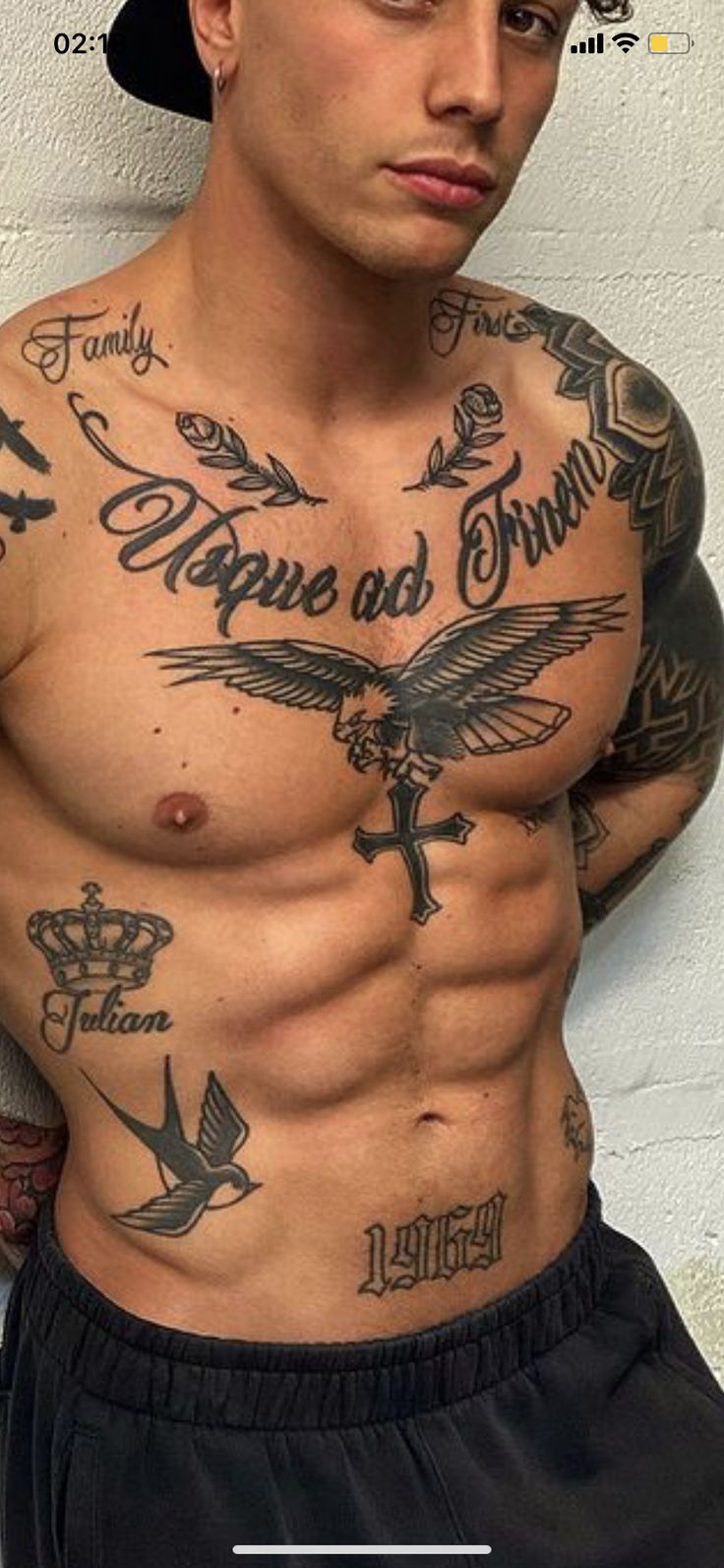 a shirtless man with tattoos on his chest and arms, leaning against a wall