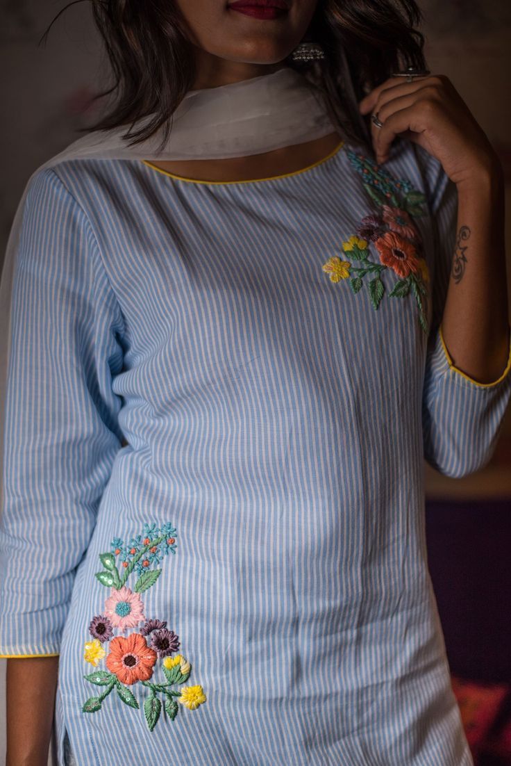 a woman wearing a blue and white striped shirt with flowers on the side, standing in front of a wall