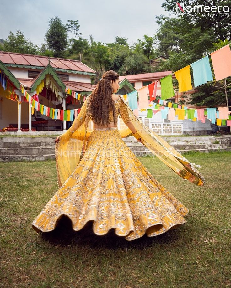 a woman in a yellow lehenga dancing on the grass with colorful flags behind her