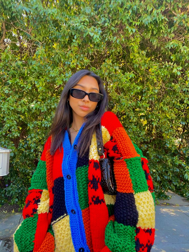 a woman standing in front of a bush wearing sunglasses and a colorful jacket with multicolored squares on it