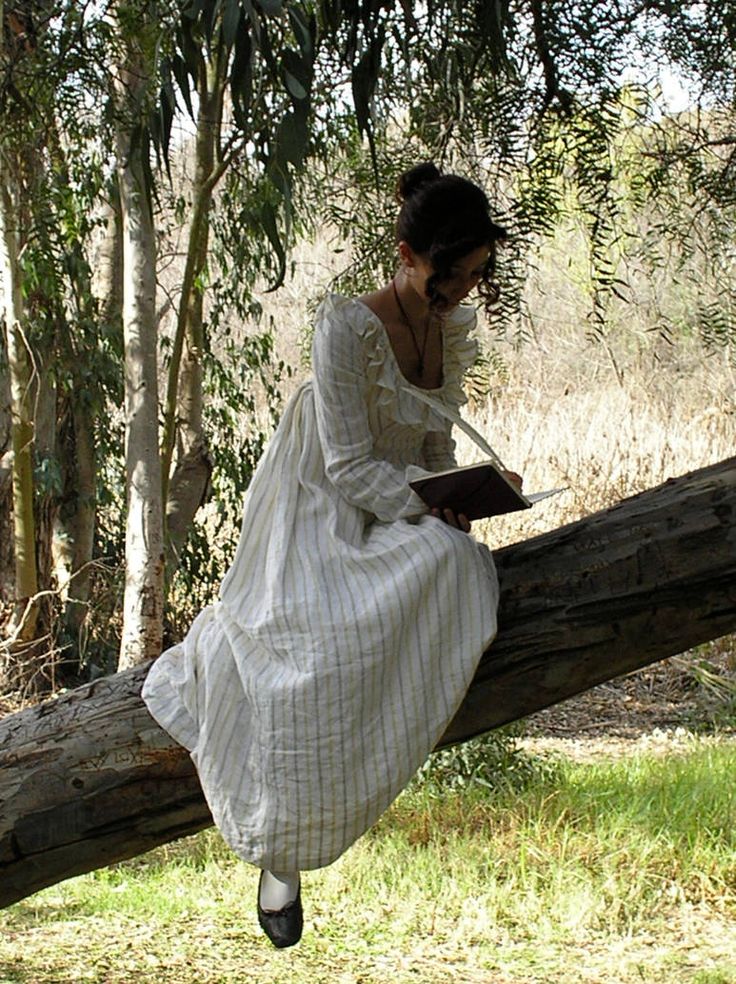 a woman wearing a white dress sitting on a log in the woods reading a book