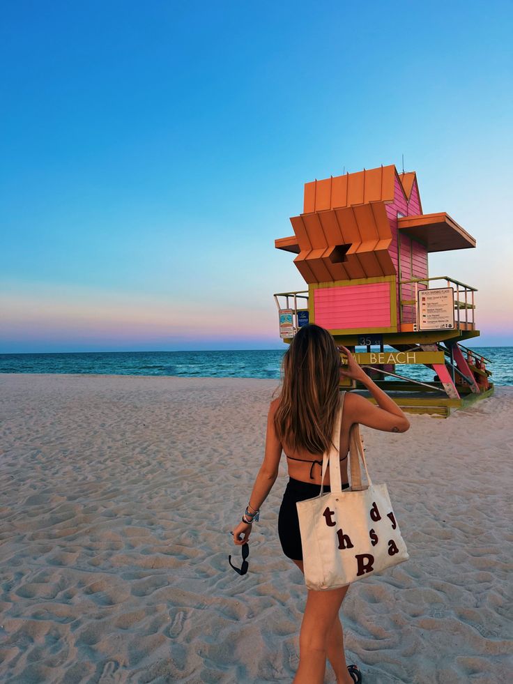 a woman is walking on the beach carrying a bag and looking into the distance with a lifeguard tower in the background