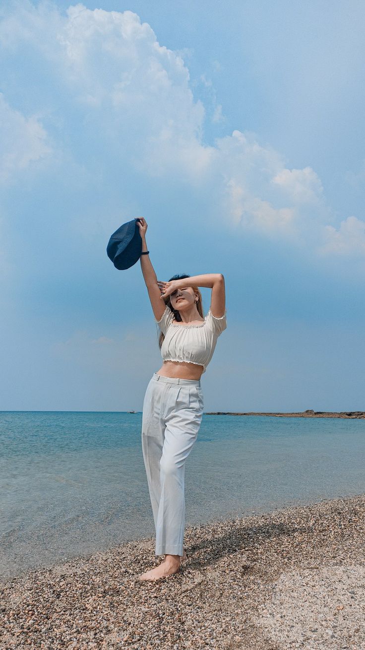 a woman standing on top of a beach next to the ocean holding a blue hat