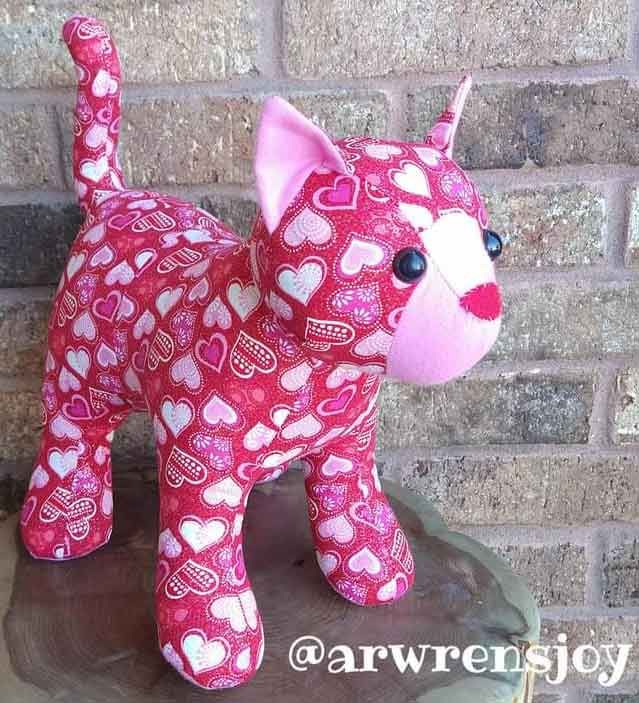 a pink stuffed cat with hearts on it's chest sitting on top of a tree stump