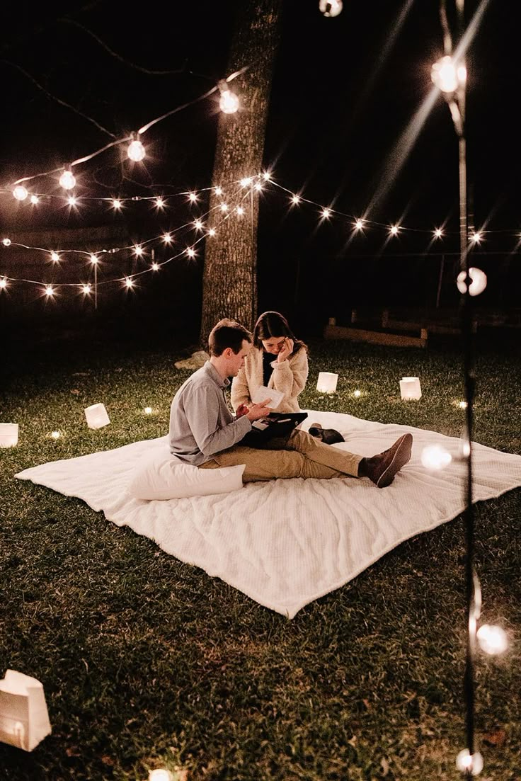 two people sitting on a blanket in the grass with lights strung around them at night