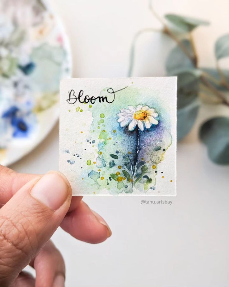 a person holding up a small card with a flower painted on it and the word bloom written in cursive writing