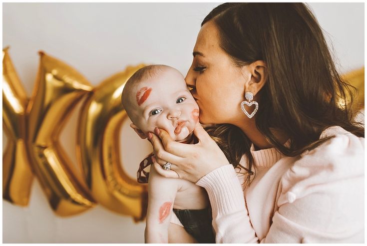 a woman holding a baby in her arms and kissing it's face with gold balloons behind her