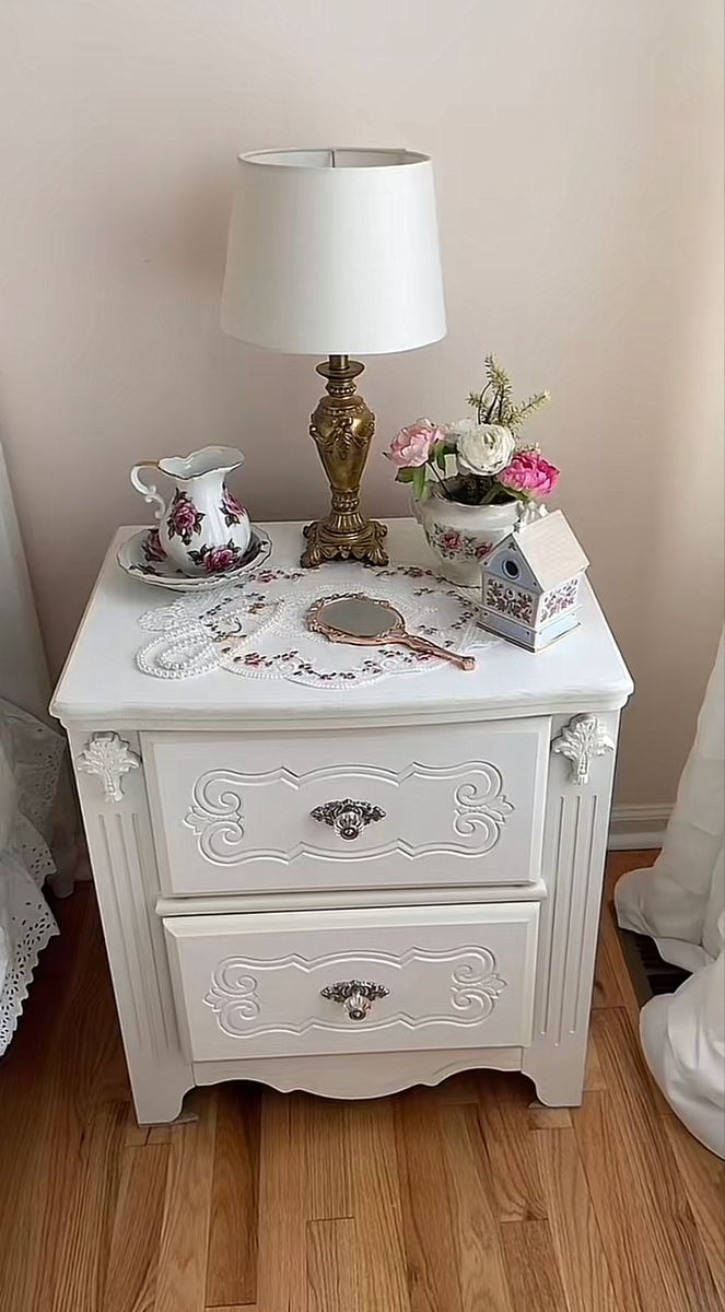 a white table topped with a lamp next to a dresser covered in jewelry and flowers