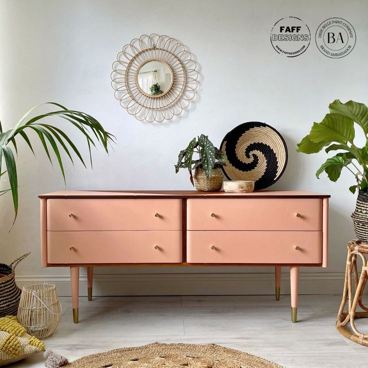 a pink dresser with two plants on top and a round mirror hanging above the drawers